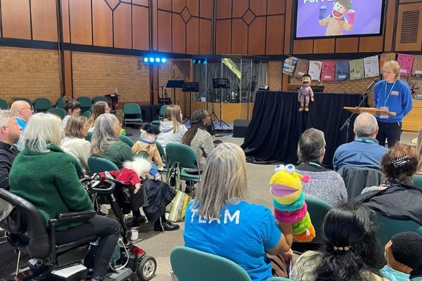 A group of adults seated in a modern church watch a presentation featuring colourful puppets, with a speaker at a lectern and a puppet displayed at the front of the room.