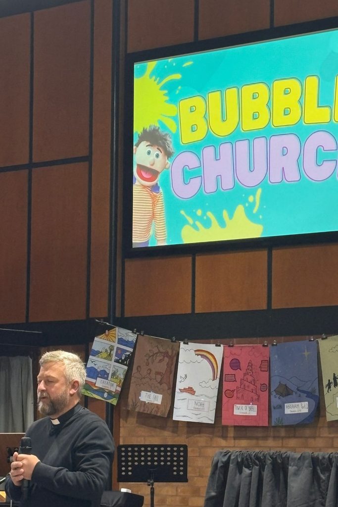 Bishop Alex holds a microphone in a church beneath a large screen displaying the Bubble Church logo and puppet mascot. Illustrated banners depicting Bible stories hang across the front of the stage area.